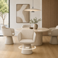 A modern dining room scene showing a round light wood table surrounded by four upholstered pedestal chairs in a warm neutral palette. A pendant light hangs above, and a vase with greenery sits on the table.