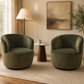 Two round barrel-style armchairs upholstered in dark olive green velvet, placed on a light beige rug in a styled living room with a side table, floor lamp, wall art, and plant. KOSSMAI
